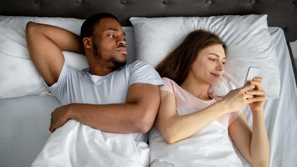 A man is lying in bed, looking suspiciously at the woman next to him who is smiling while looking at her phone.
