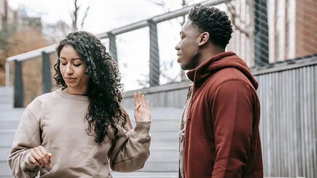 A woman with long curly hair is holding up her hand toward a man in a red hoodie outdoors.