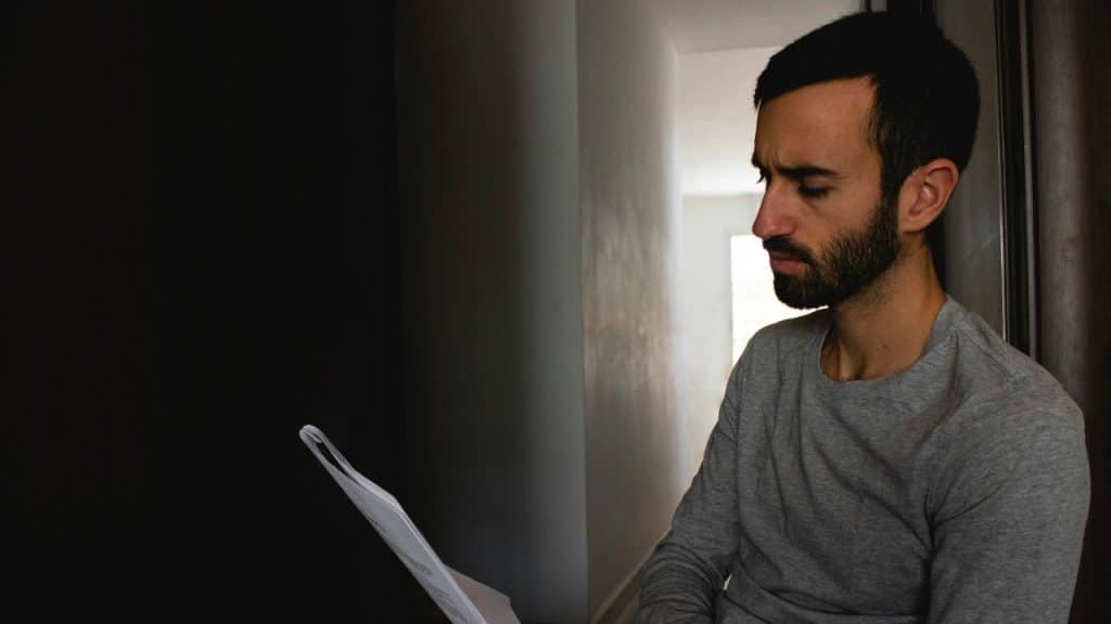 A man with a beard leans against a wall in a hallway while reading papers.