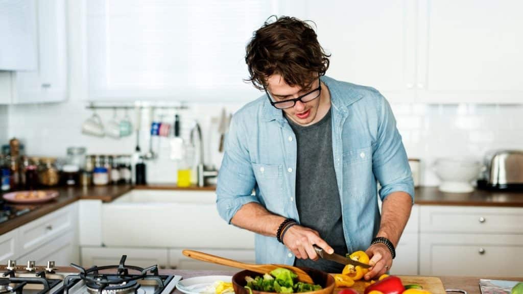 A man with glasses and a denim shirt chops yellow peppers in a white kitchen.