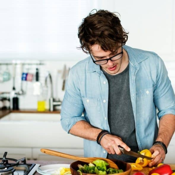 A man with glasses and a denim shirt chops yellow peppers in a white kitchen.