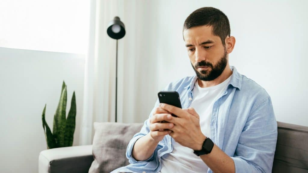 A man with a beard sits on a grey sofa looking down at his smartphone.