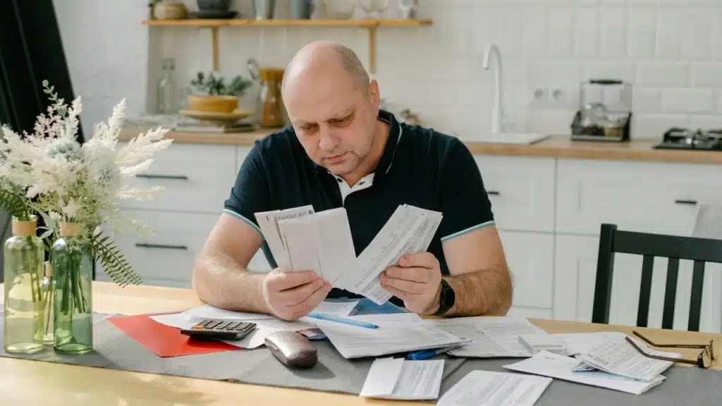 A man sits at a wooden table reviewing stacks of white papers and a calculator.