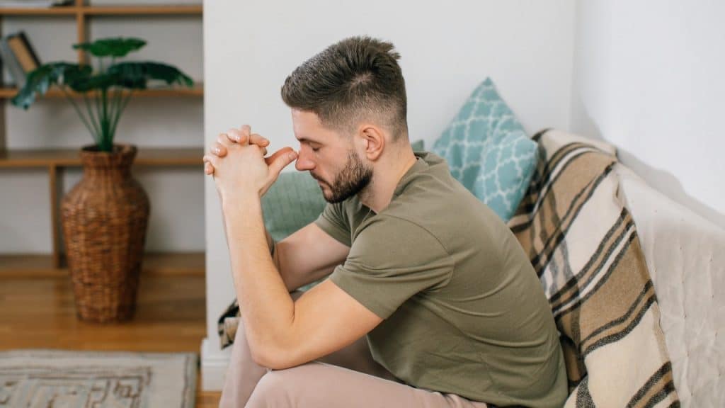 A man with a beard sits on a sofa with his hands clasped together near his face.