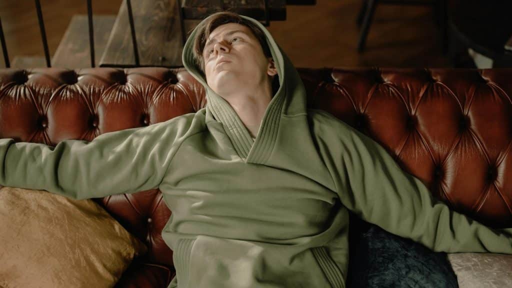 A person wearing a green hooded sweatshirt leans back on a brown tufted leather sofa.