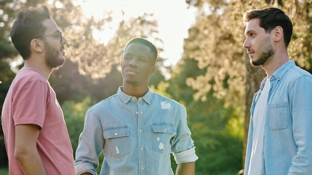 Three men stand outdoors in a park during sunset, engaged in a conversation together.