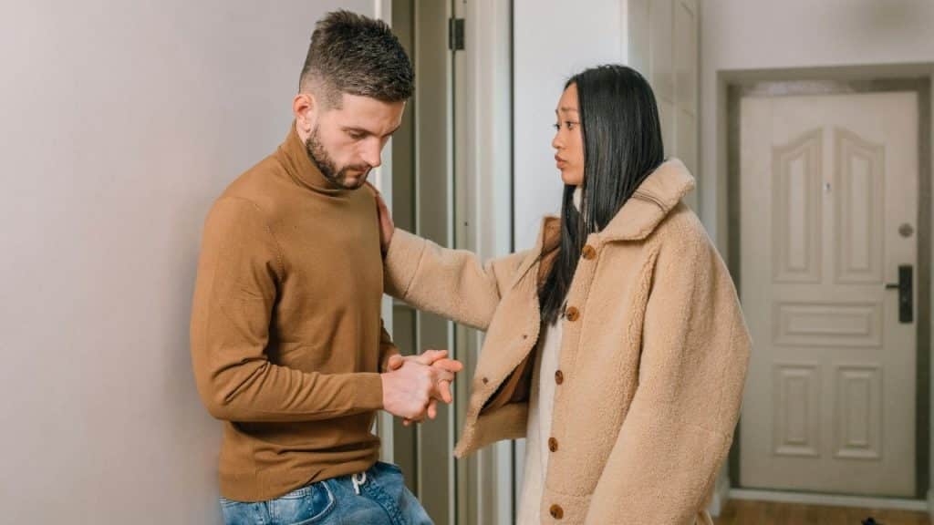 A woman in a tan coat places her hand on a man’s shoulder in a hallway.
