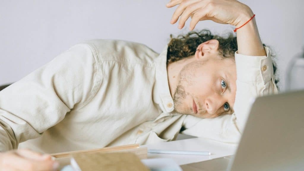 A person with curly hair rests their head on their arm on a desk with a laptop.