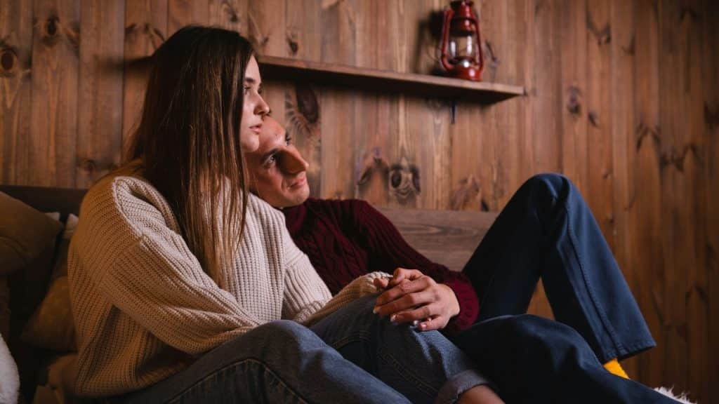A couple sits closely together on a couch against a rustic wooden paneled wall.