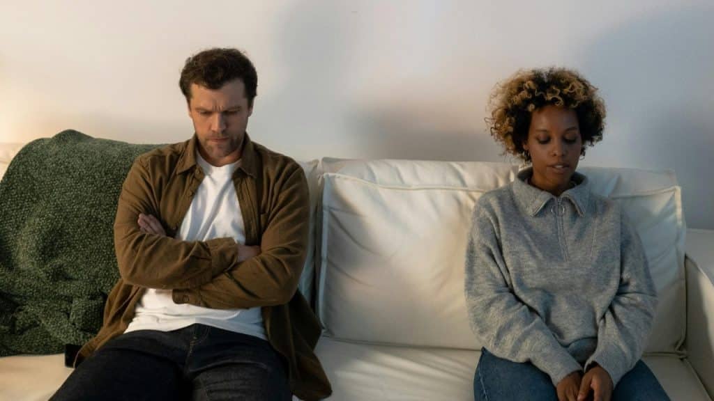 A man with crossed arms and a woman sit apart on a white sofa.