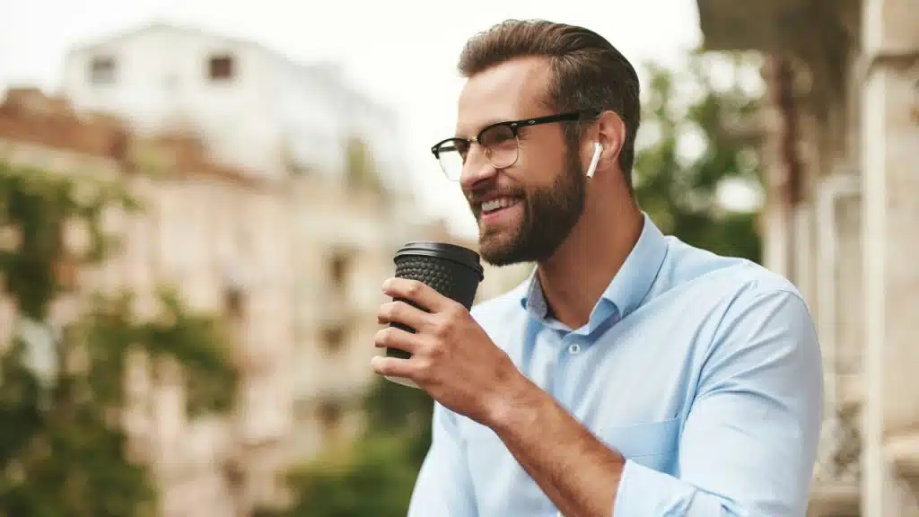 A smiling man with glasses and wireless earbuds holds a black coffee cup outdoors.