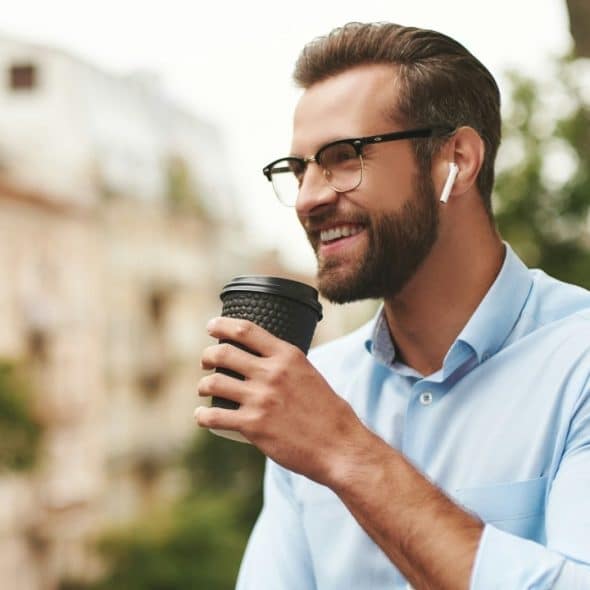 A smiling man with glasses and wireless earbuds holds a black coffee cup outdoors.