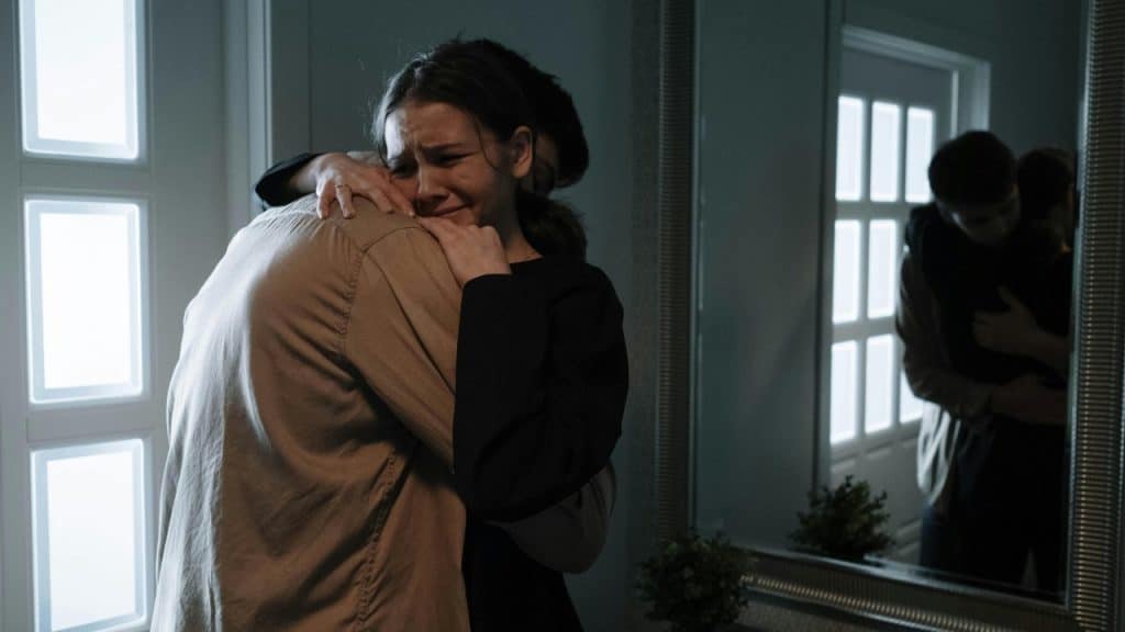 A woman cries while embracing a man in a dimly lit room with a mirror.