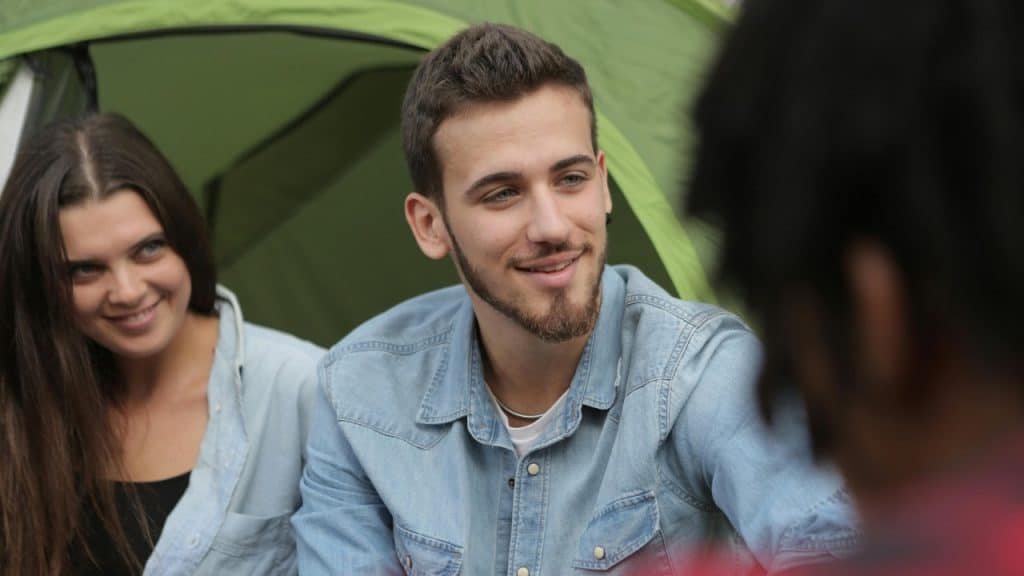 A man with a beard smiles while sitting with others in front of a tent.