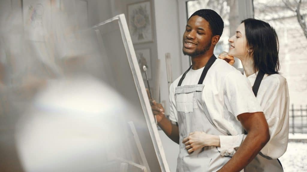 A woman hugs a man from behind while he holds a paintbrush near a canvas.