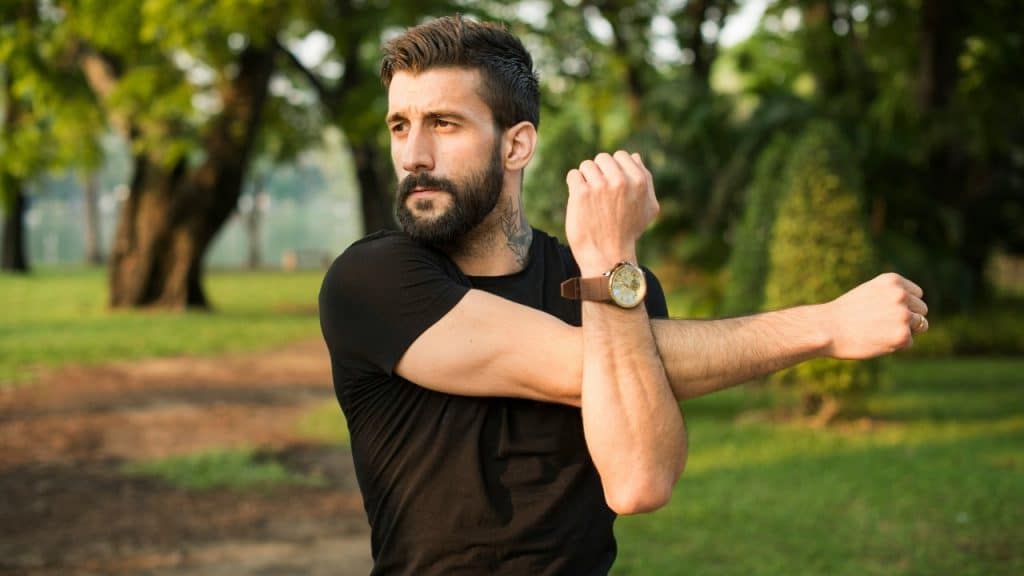 A bearded man in a black shirt stretches his arms while standing in a park.
