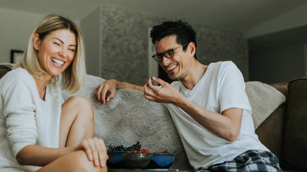 A man and woman sit on a couch laughing while eating snacks from small bowls.