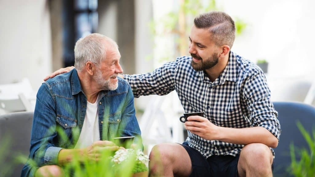 Two men sit outside holding coffee cups, with the younger man's hand on the older's shoulder.