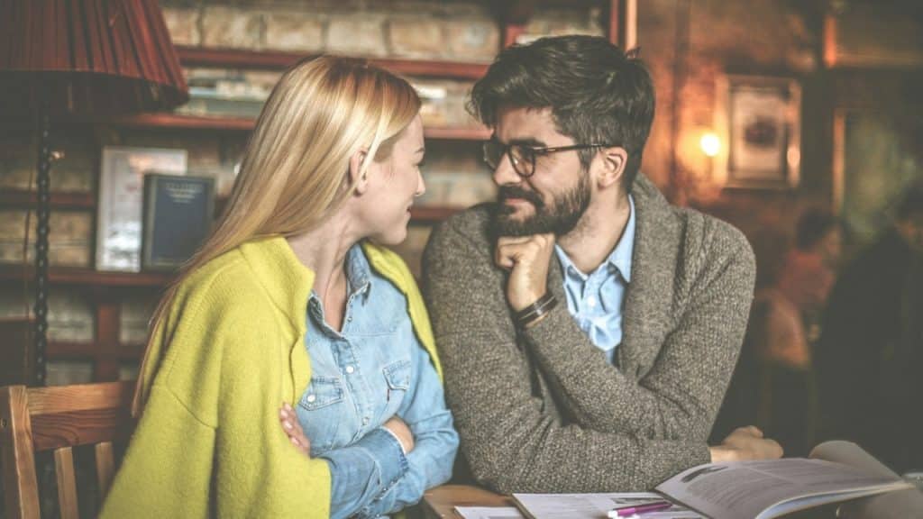 A man and woman sit at a table with an open book, looking at each other.