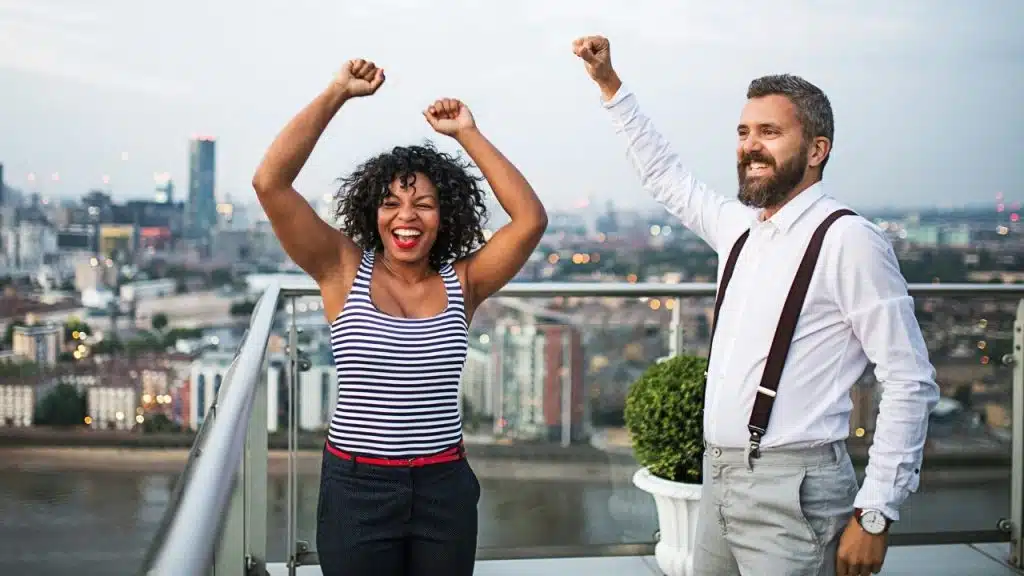 A man and woman cheer with raised fists on a balcony overlooking a city skyline.