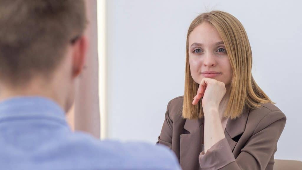 A woman in a brown blazer sits at a table while facing a blurry person.