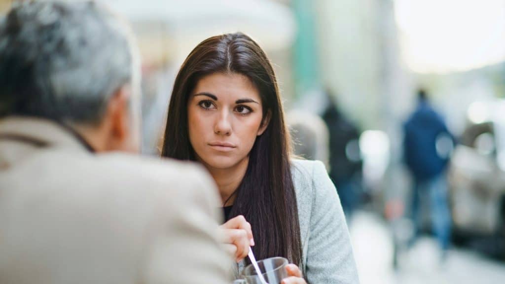 A woman with long dark hair looks at a blurry man across a cafe table.