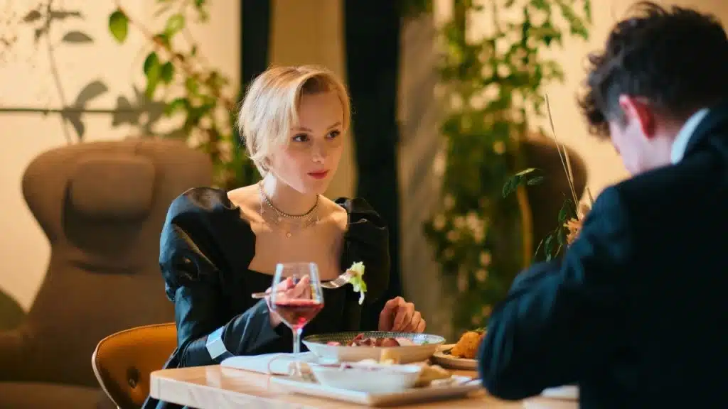 A blonde woman in a black puff-sleeve dress eats dinner across from a man.