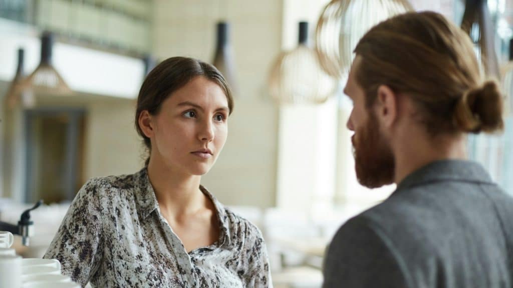 A woman in a patterned shirt looks directly at a man with a hair bun.