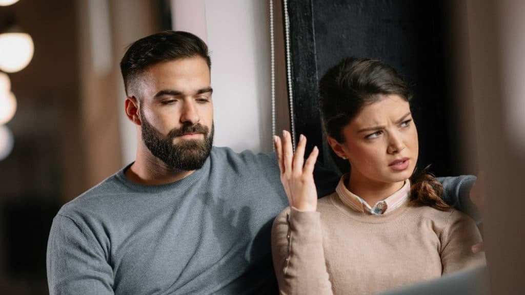 A woman with a raised hand looks away while a bearded man sits beside her.