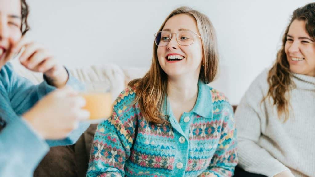 A woman wearing glasses and a blue patterned cardigan laughs while sitting with two others.