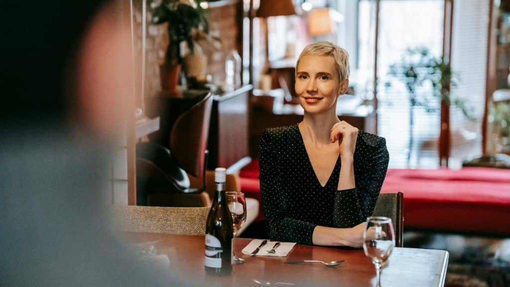 A woman with short blonde hair sits at a table with a bottle and glasses.
