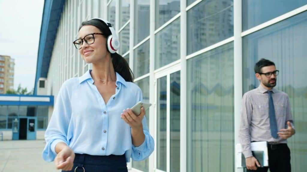 A woman wearing headphones and glasses holds a phone while walking past a glass building.