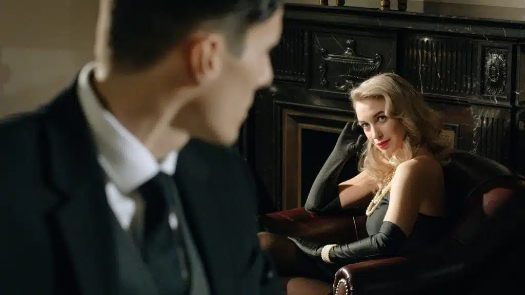 A woman in a black dress and gloves sits in a leather chair near a fireplace.