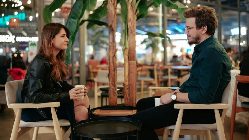 A man and woman sit in chairs facing each other while holding mugs in a cafe.
