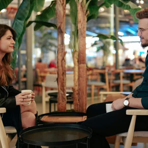 A man and woman sit in chairs facing each other while holding mugs in a cafe.