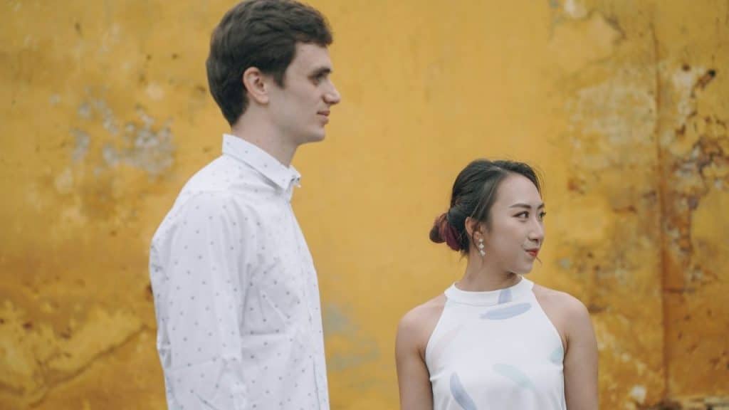 A man and a woman stand side-by-side in profile against a textured yellow wall.
