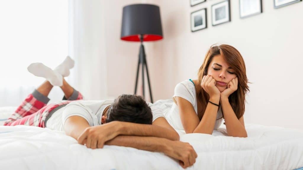 A man and woman lie on a bed looking away from each other with downcast expressions.