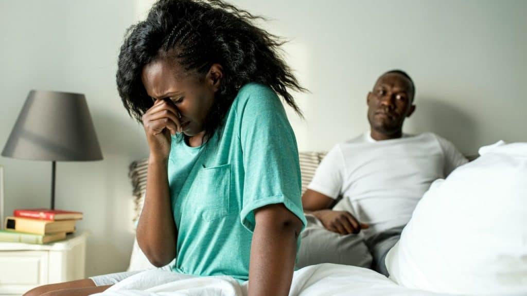 A woman sits on a bed covering her face while a man sits in the background.