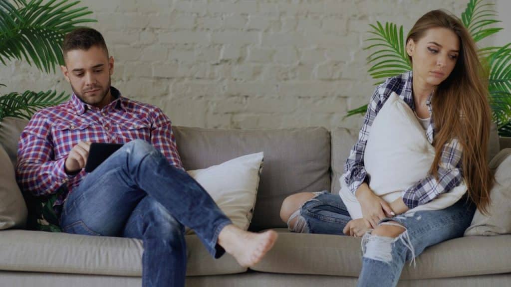 A man and woman sit on a sofa looking at a tablet and away, respectively.