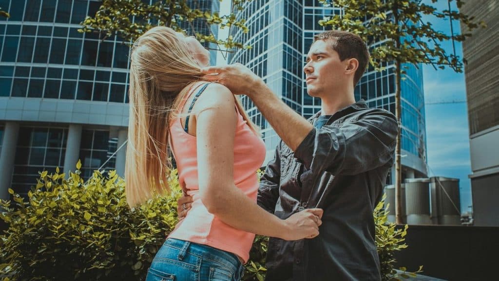 A man and woman stand close together outdoors in front of a modern glass building.