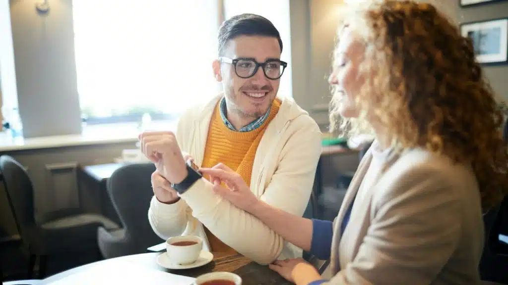 A man in a yellow sweater shows a watch to a woman at a table.