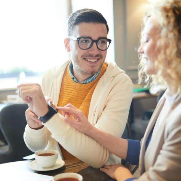 A man in a yellow sweater shows a watch to a woman at a table.
