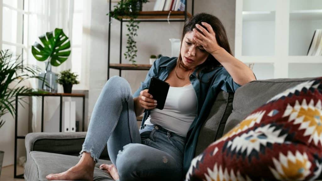A woman sits on a gray sofa holding a phone while touching her forehead with one hand.