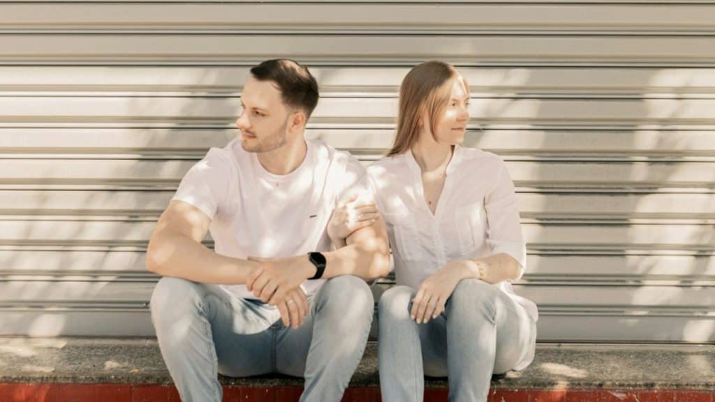 A man and woman sit side-by-side on a ledge in front of a metal shutter.