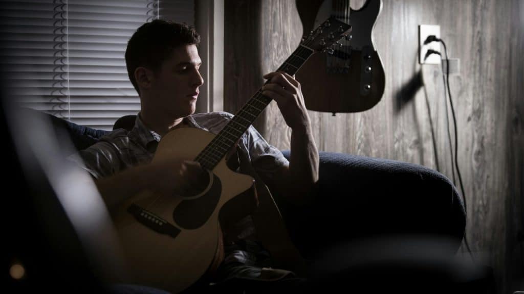 A man plays an acoustic guitar in a dimly lit room with another guitar hanging.