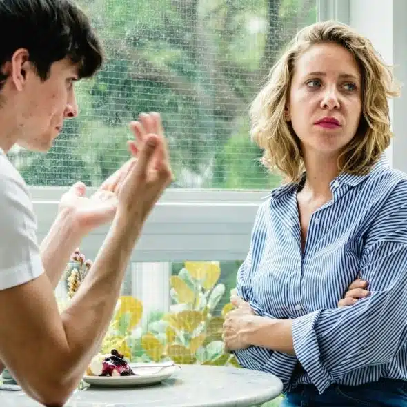 A man in a white shirt gestures while a woman with crossed arms looks away.