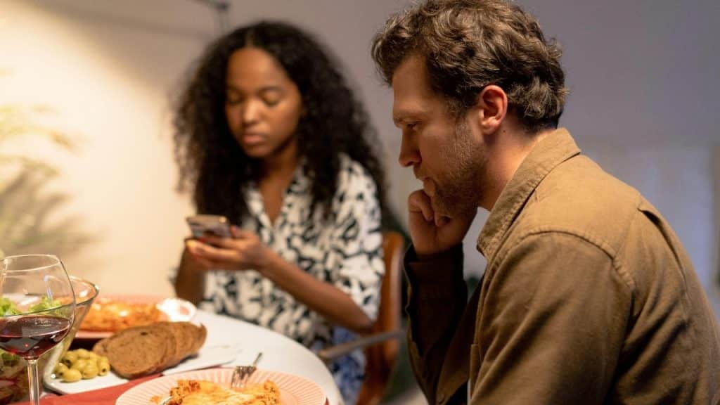 A man looks down at a table while a woman sits behind him using a phone.