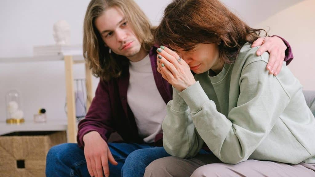 A man sits with his hand on a woman's shoulder as she covers her face.