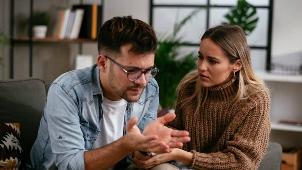 A man with glasses gestures with open palms while a woman in a sweater watches.