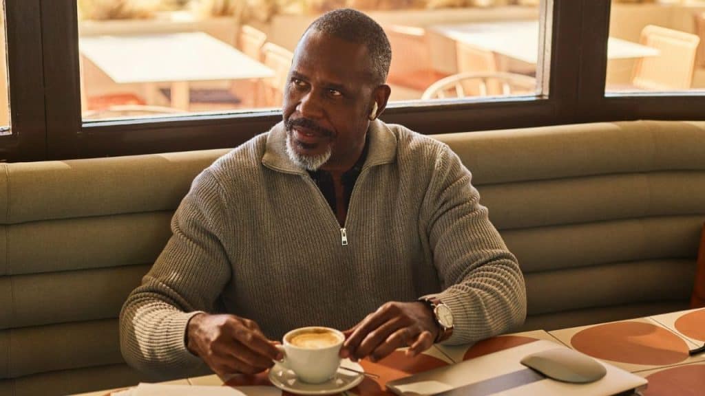 A man with a grey beard sits at a table holding a white cup of coffee.